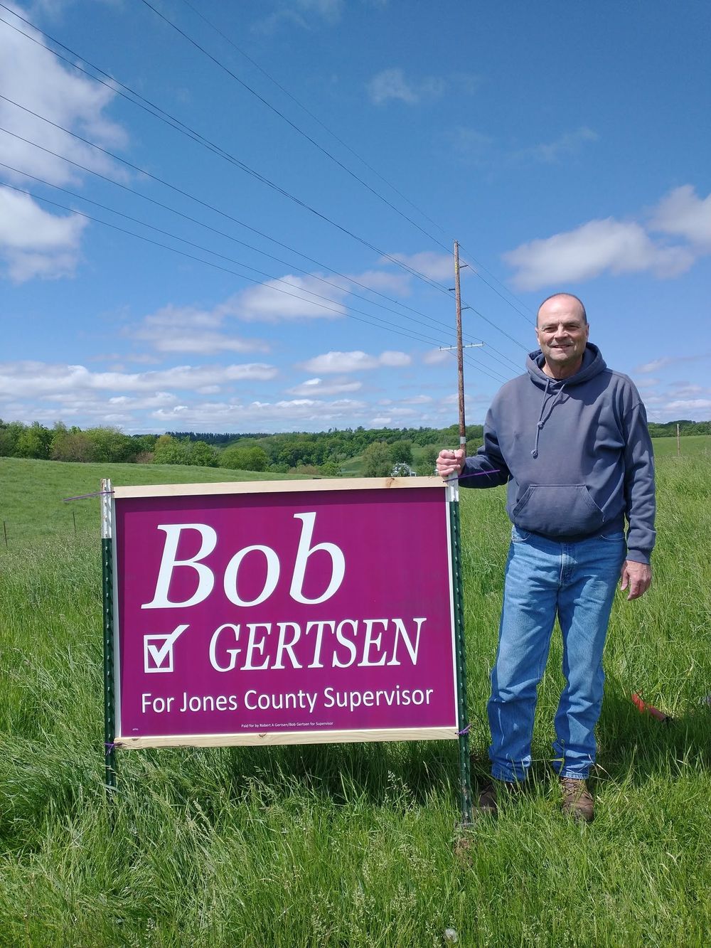 Bob Gertsen standing beside a large campaign sign in a green field